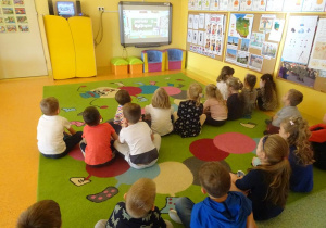 Grupa dzieci siedzi na dywanie, ogląda film edukacyjny na tablicy interaktywnej.